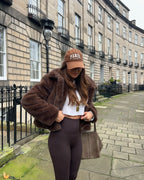 Woman wearing a brown faux fur cropped jacket over a white crop top and brown leggings, holding her hands to the jacket.
