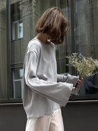 Side view of a woman in a grey oversized knit sweater with bell sleeves and a white skirt, holding dried flowers.