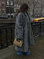 Woman wearing a long grey faux fur coat and blue jeans, standing by a canal in a city.