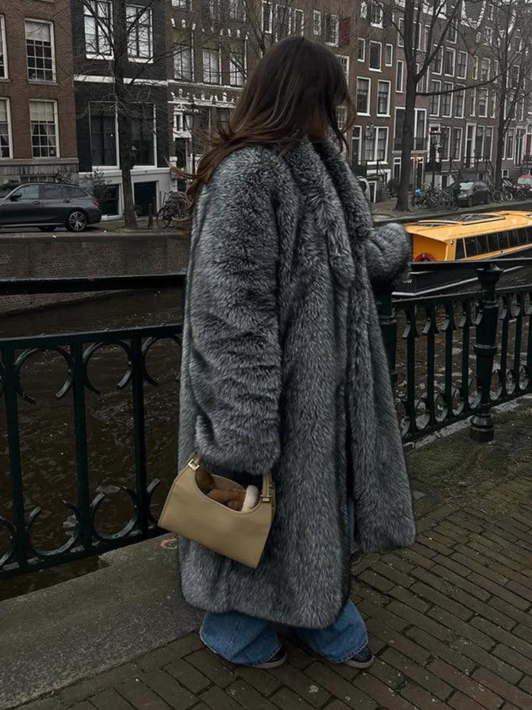 Woman wearing a long grey faux fur coat and blue jeans, standing by a canal in a city.