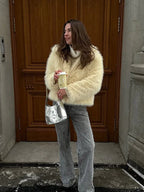 Woman in a cream faux fur jacket, grey jeans, and a silver handbag, holding a coffee cup.