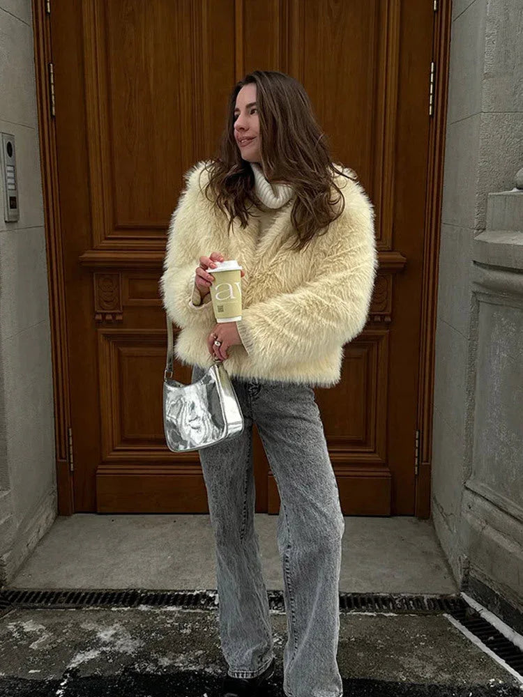 Woman in a cream faux fur jacket, grey jeans, and a silver handbag, holding a coffee cup.