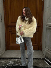Woman in a cream faux fur jacket, grey jeans, and a silver handbag, holding a coffee cup.