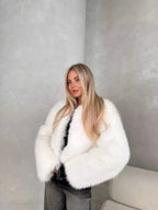 Woman in a white fluffy faux fur cropped jacket and dark wash jeans, posing with hands tucked into the jacket.