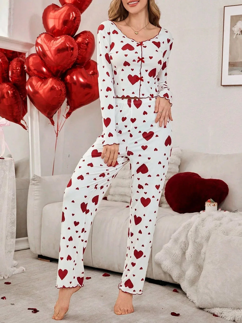 Woman wearing a white long-sleeved pajama top and pants with a red heart print, standing with a leg bent.