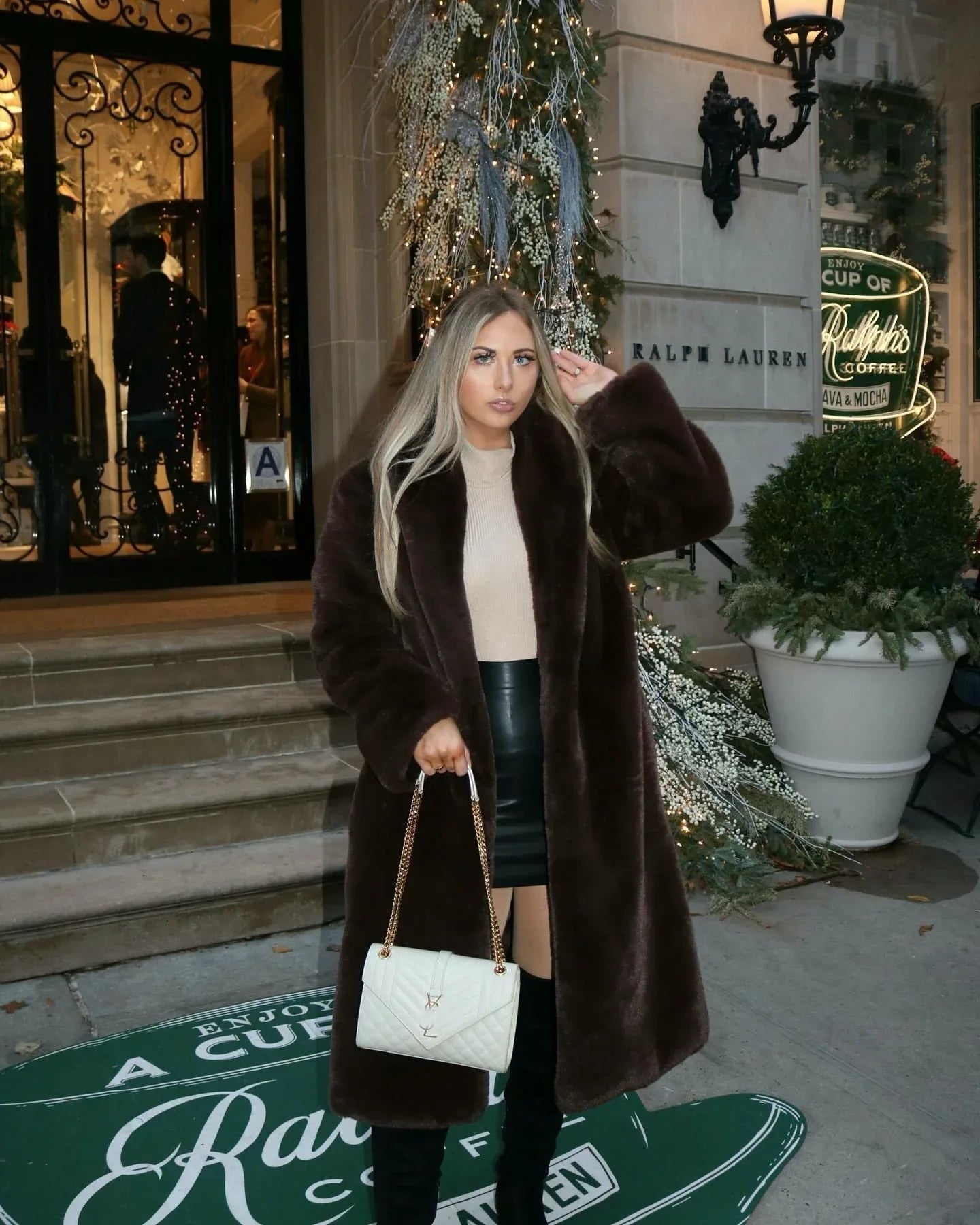 Woman wearing a long brown faux fur coat over a beige turtleneck and black faux leather mini skirt, holding a white quilted handbag. Main image