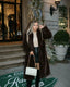 Woman wearing a long brown faux fur coat over a beige turtleneck and black faux leather mini skirt, holding a white quilted handbag.