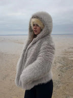 Woman standing on a beach, wearing a grey faux fur hooded coat