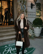 Woman looking down, wearing a long brown faux fur coat, beige turtleneck, and black faux leather mini skirt, holding a white quilted handbag.