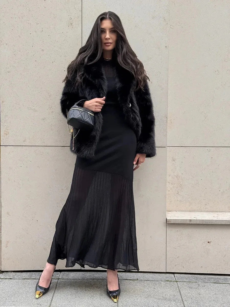 Woman wearing a black faux fur jacket over a black dress and heels, posing against a wall.