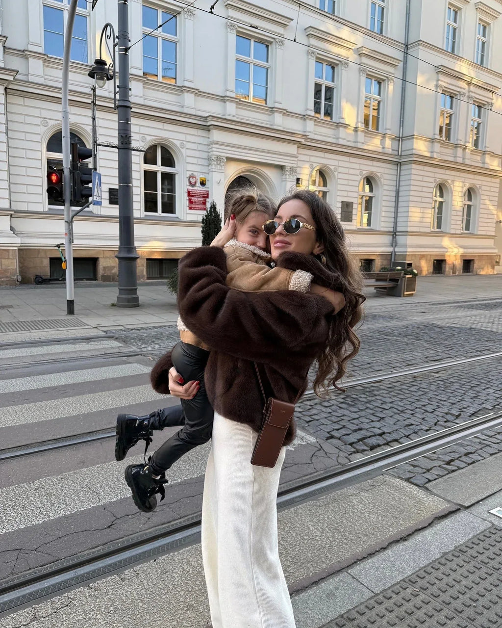 Woman in a brown faux fur jacket holding a child in her arms on a street crossing.