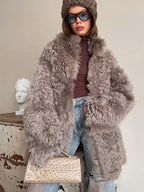 Front view of a woman in a gray curly faux shearling coat, brown turtleneck, ripped blue jeans, and a brown beanie, holding a beige clutch bag.