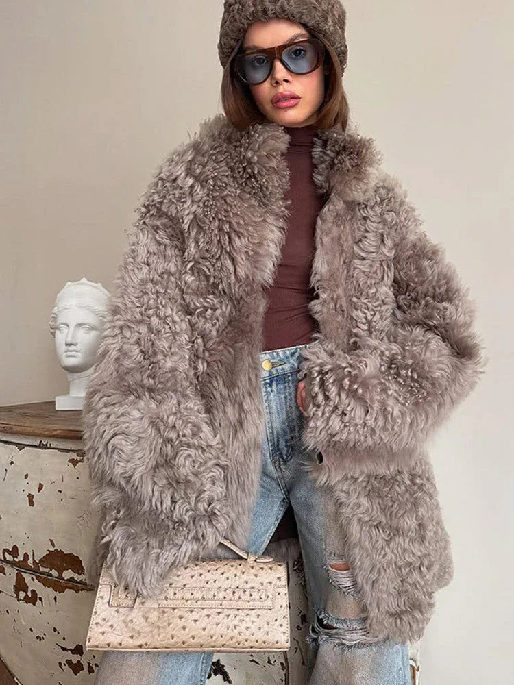 Front view of a woman in a gray curly faux shearling coat, brown turtleneck, ripped blue jeans, and a brown beanie, holding a beige clutch bag.