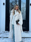 Woman wearing a cream faux fur long coat with a chevron pattern, white turtleneck sweater, and blue jeans, holding a dark green handbag, standing in front of a building with snow.