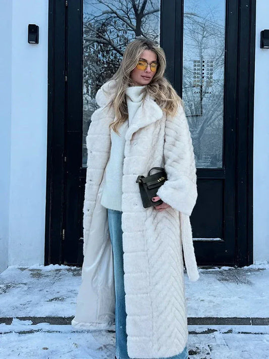 Woman wearing a cream faux fur long coat with a chevron pattern, white turtleneck sweater, and blue jeans, holding a dark green handbag, standing in front of a building with snow.
