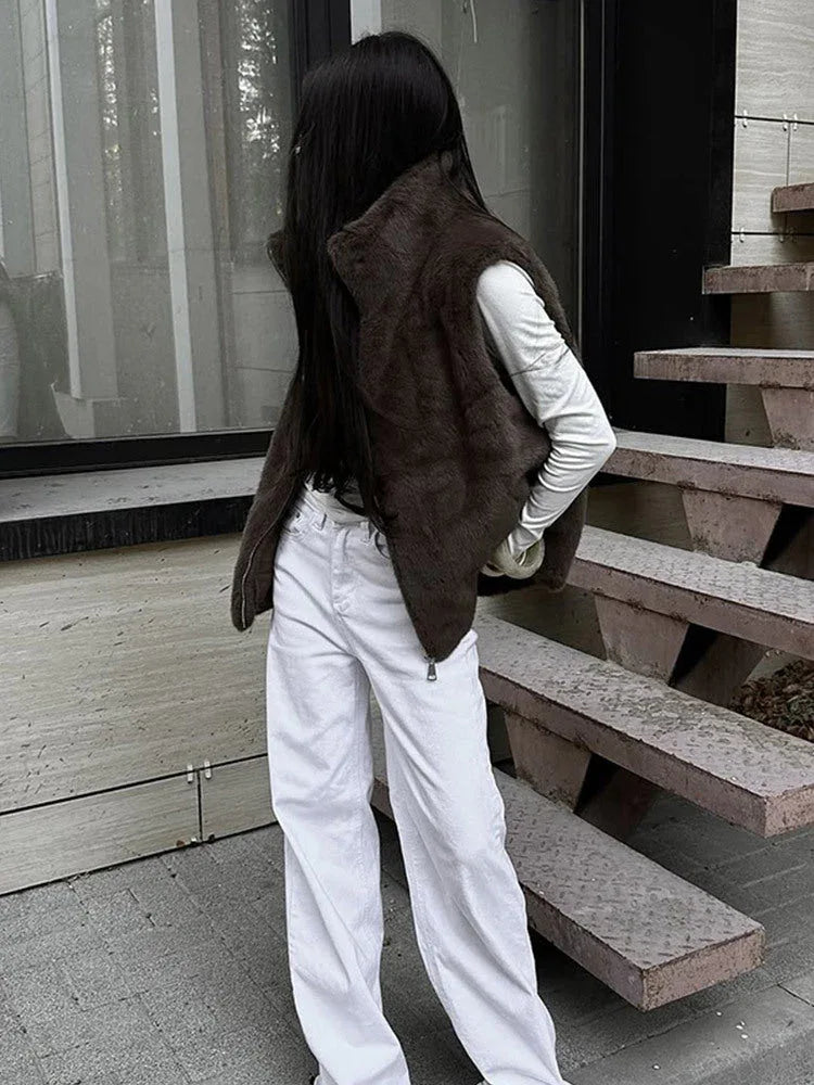 Woman wearing a brown faux fur vest over a white long-sleeved shirt and white pants, standing outdoors. Main image