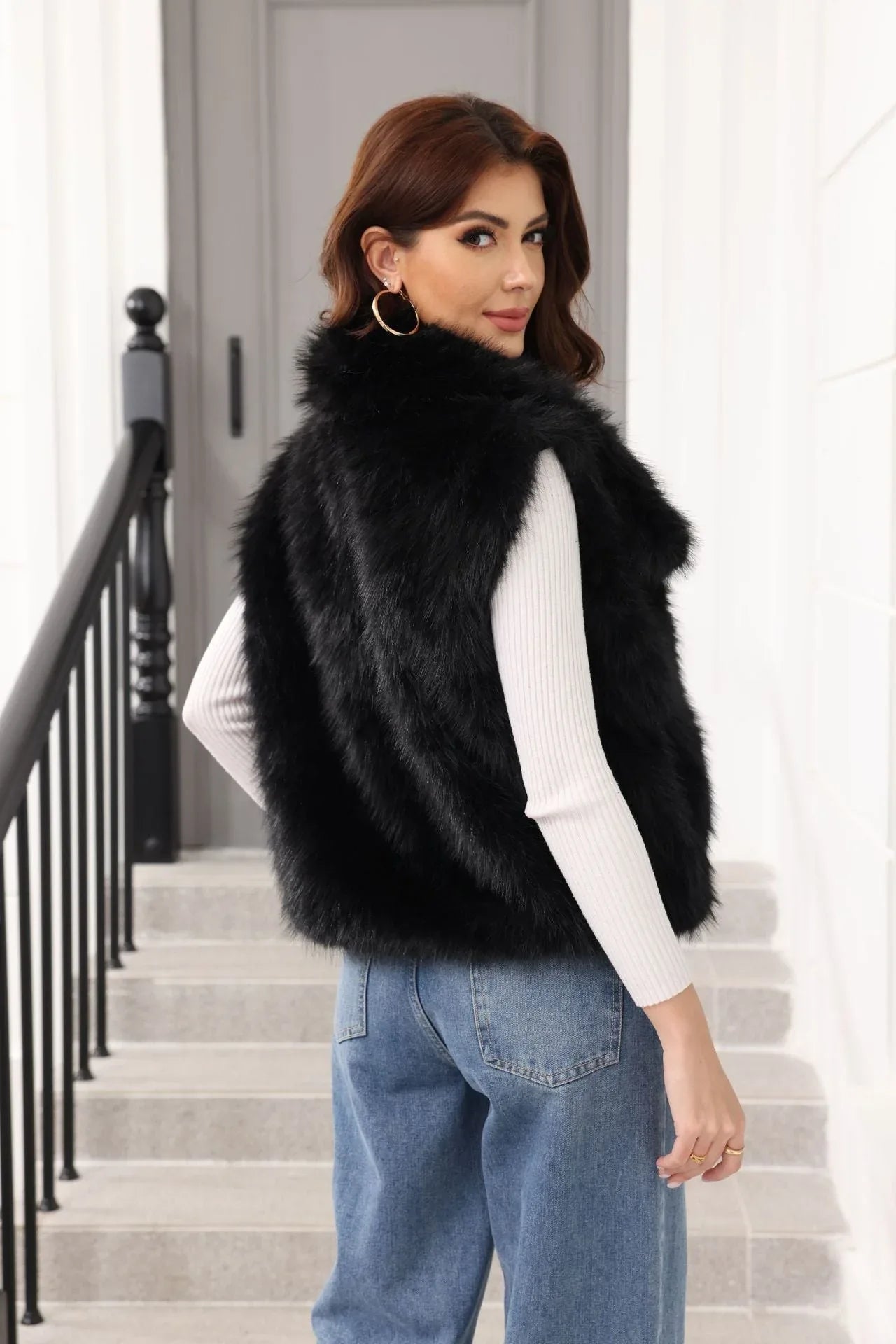 Woman wearing a black faux fur vest over a white long-sleeved shirt and blue jeans, looking over her shoulder