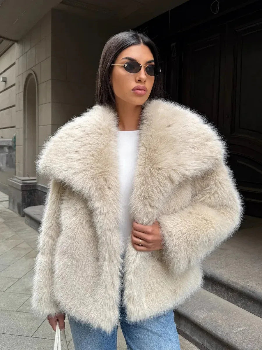 Woman in a cream-colored faux fur jacket, white top, blue jeans, and sunglasses, holding a white handbag.