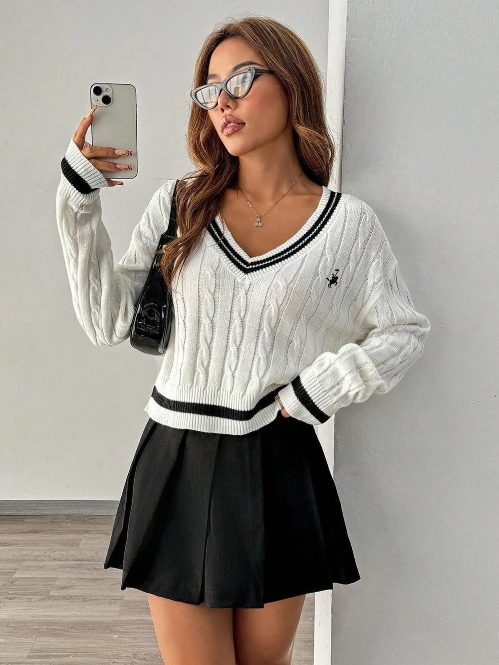 Woman taking a selfie while wearing a white cable knit cropped sweater with black trim and a black pleated skirt.