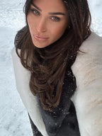 Close-up of a woman's face, wearing a black and white faux fur coat and sunglasses, with snow in the background.