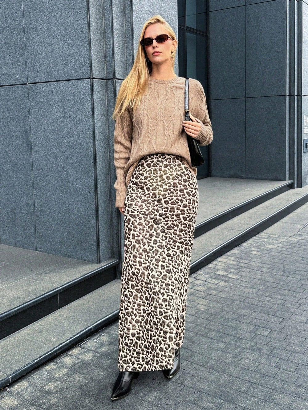 Full body shot of a woman in a tan cable knit sweater and leopard print maxi skirt, with a black handbag.