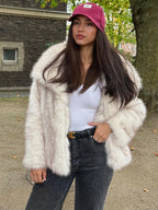 Woman in a cream faux fur jacket, white tank top, and dark jeans, outdoors.
