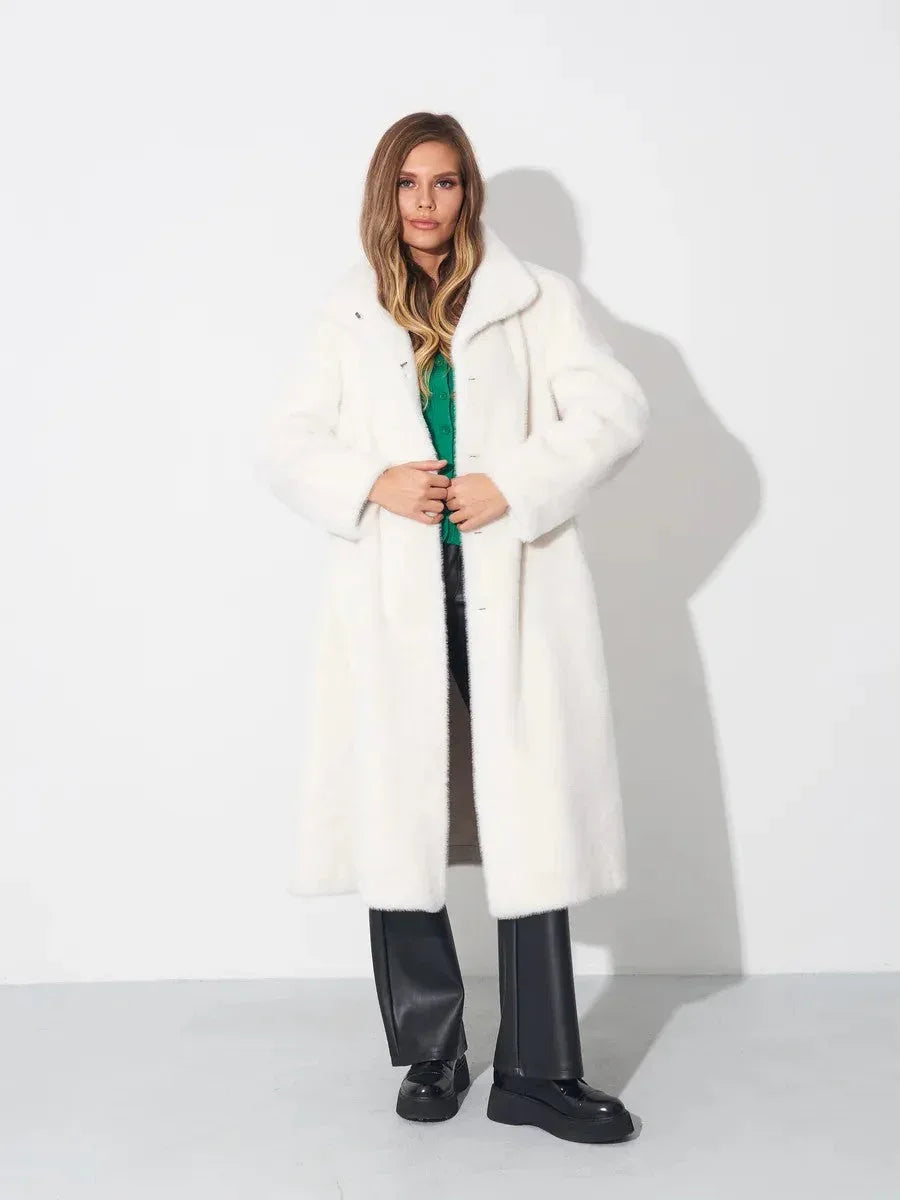 Woman wearing a long white faux fur coat, green top, and black pants, standing against a white wall.
