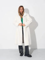 Woman wearing a long white faux fur coat, green top, and black pants, standing against a white wall.