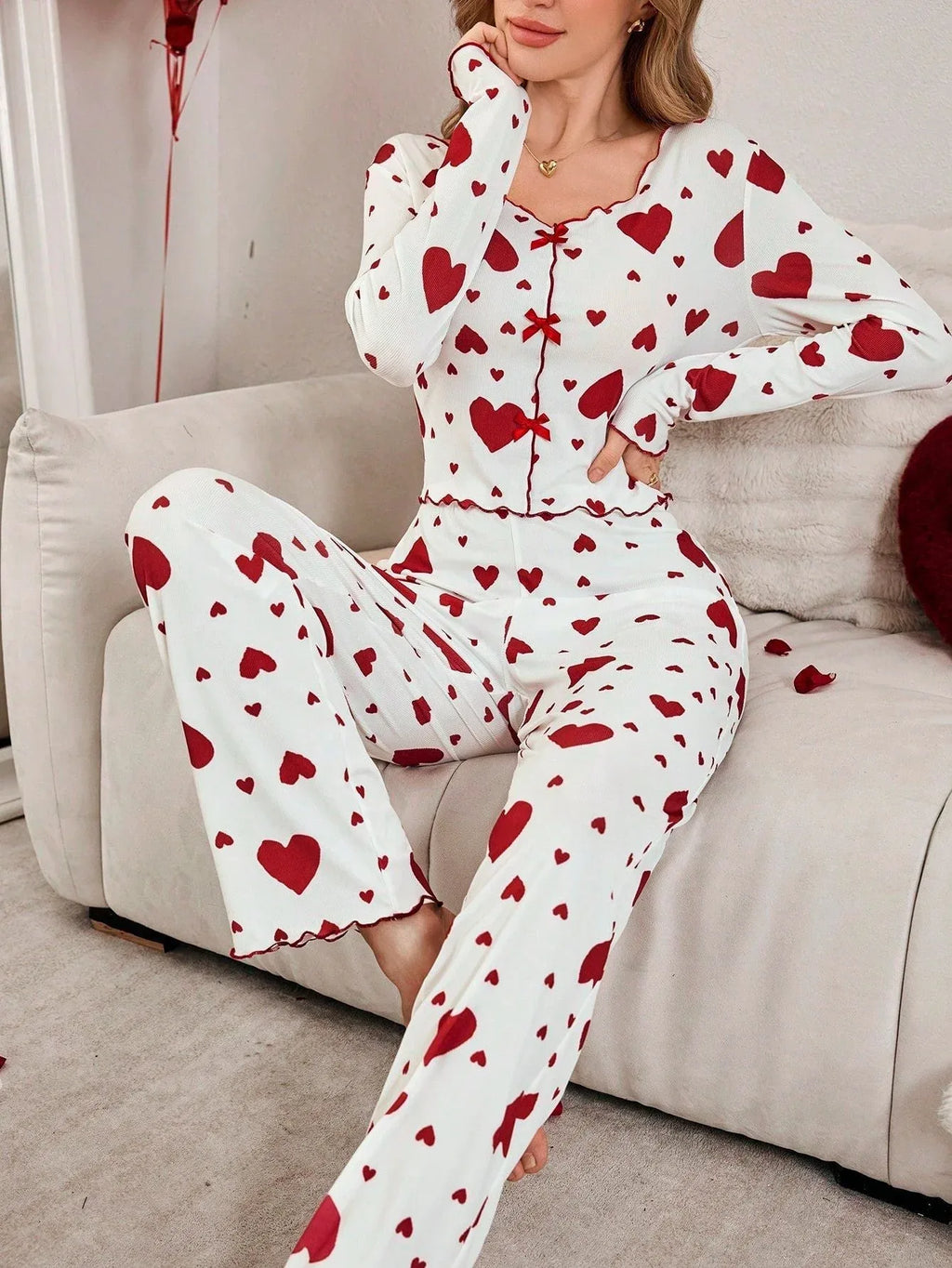 Woman wearing a white long-sleeved pajama top and pants with a red heart print, sitting on a couch with her hand on her chin.