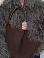 Close-up of a woman's hands holding a brown tote bag, with a shaggy grey faux fur jacket visible.