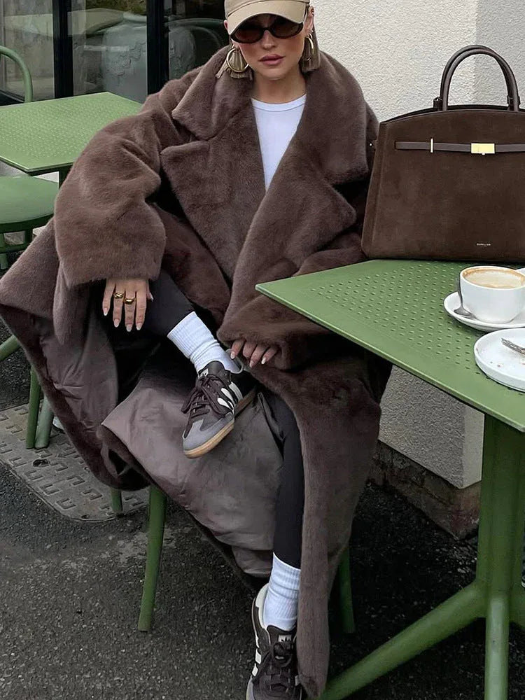 A woman sits at a green outdoor cafe table, wearing a brown faux fur coat and holding a coffee cup.
