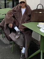 A woman sits at a green outdoor cafe table, wearing a brown faux fur coat and holding a coffee cup.