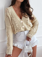 Woman wearing a cream-colored, open-knit, cropped cardigan with a V-neck and button-up front, paired with white denim shorts.