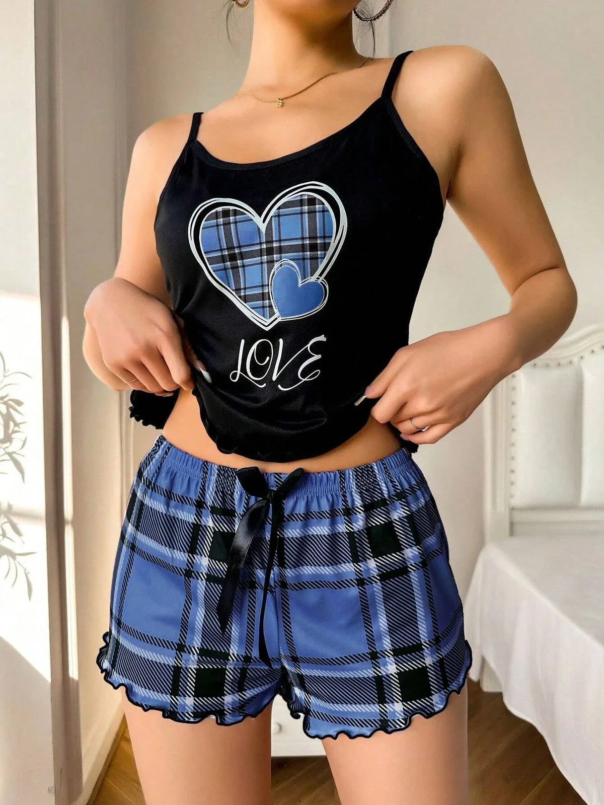 Woman standing and adjusting the hem of her black camisole with a blue plaid heart graphic, revealing blue plaid pajama shorts with a drawstring waist and ruffle trim.