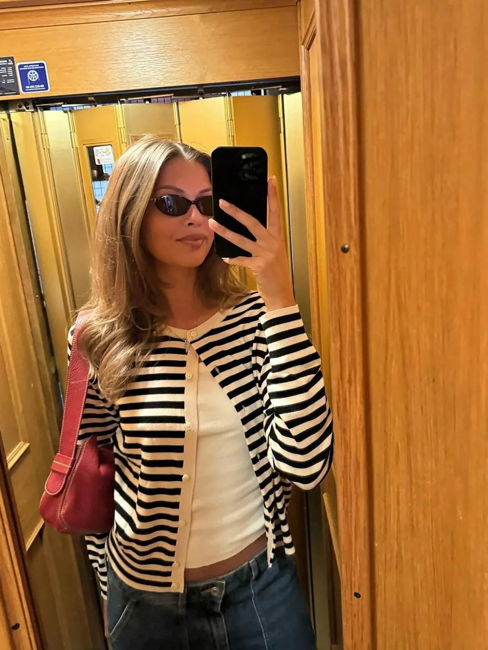 Woman in a black and white striped cardigan and blue jeans taking a mirror selfie in an elevator.