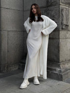 Woman wearing a long white fuzzy knit cardigan over a white ribbed dress and white boots, posing against a stone building.
