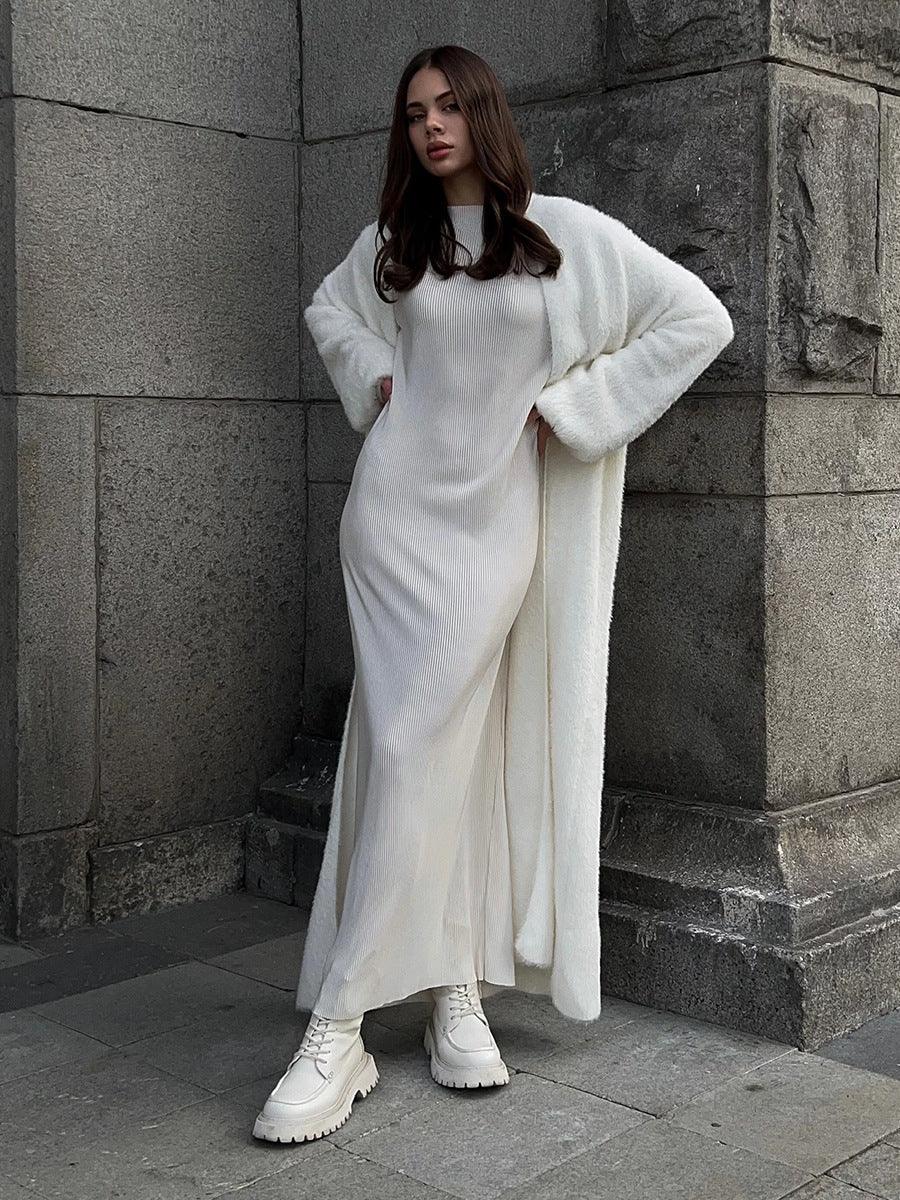 Woman wearing a long white fuzzy knit cardigan over a white ribbed dress and white boots, posing against a stone building.