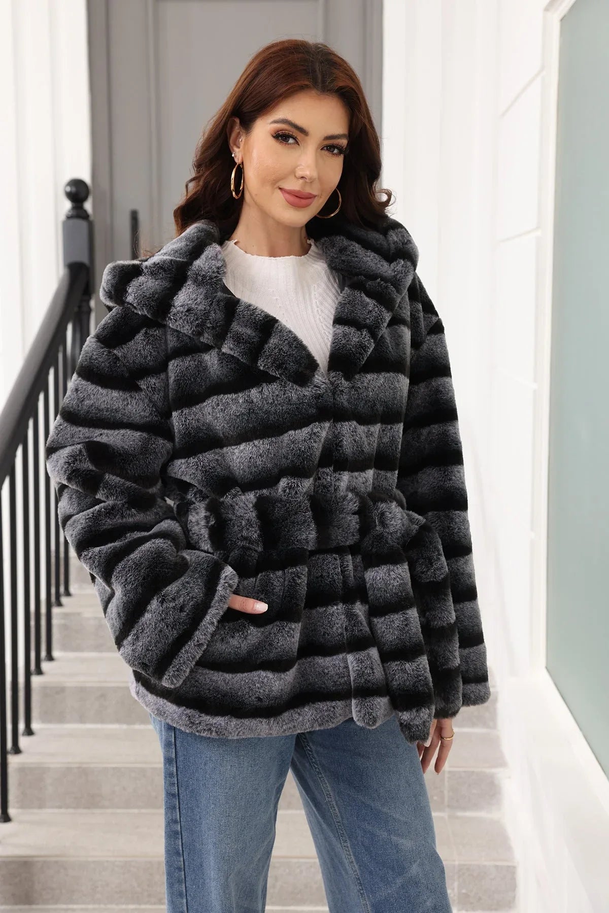 Woman wearing a hooded faux fur jacket with horizontal black and grey stripes and a belted waist.