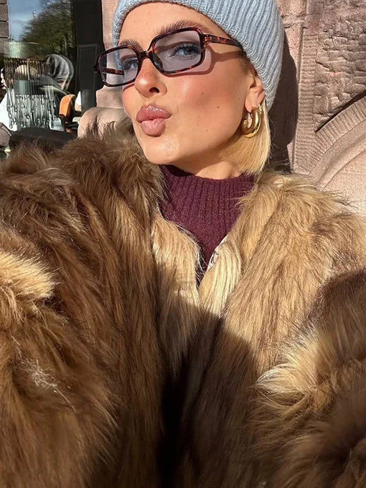 Close-up of a woman wearing a tan faux fur jacket, blue beanie, and glasses, pouting.