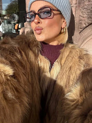 Close-up of a woman wearing a tan faux fur jacket, blue beanie, and glasses, pouting.