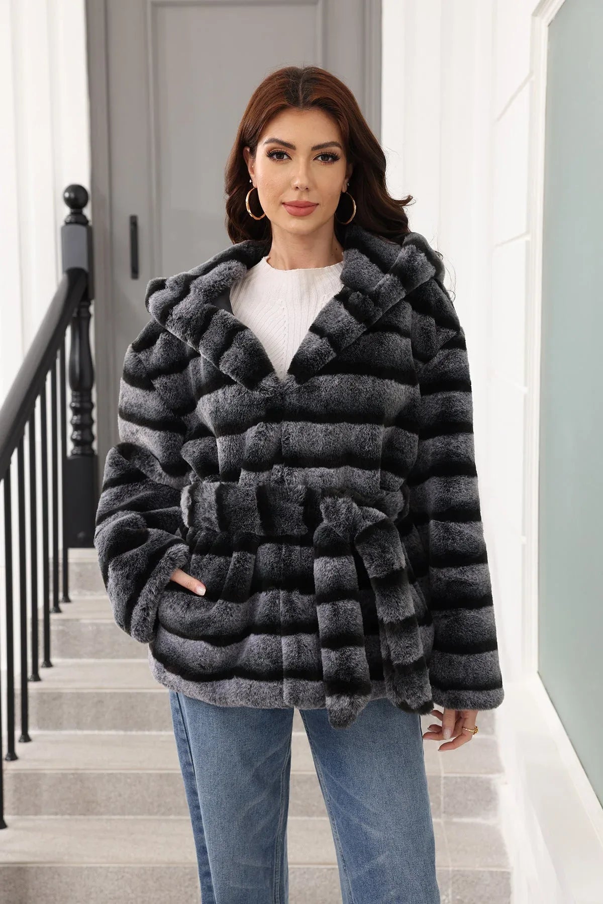Woman wearing a hooded faux fur jacket with horizontal black and grey stripes and a belted waist, standing on stairs.
