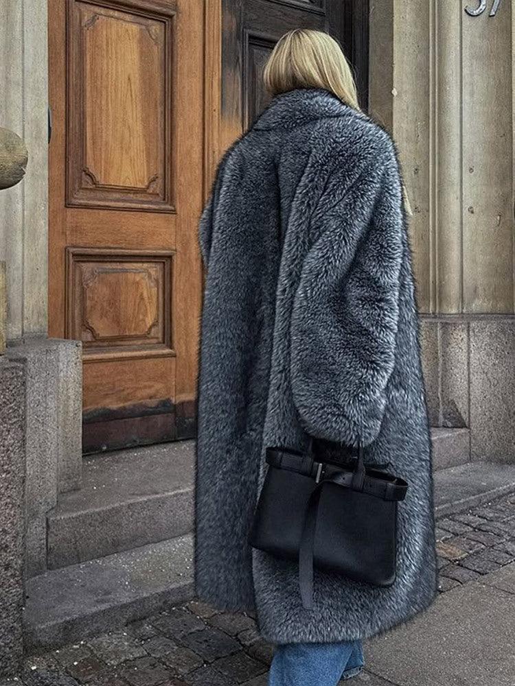 Woman wearing a long grey faux fur coat and blue jeans, standing outside a wooden door.