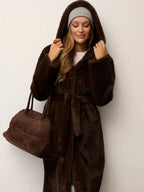 Woman in a brown hooded faux fur coat, grey beanie, and holding a brown suede bag.