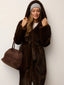 Woman in a brown hooded faux fur coat, grey beanie, and holding a brown suede bag.