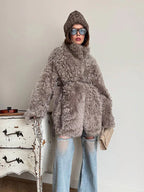 Woman wearing a gray curly faux shearling coat with a belt, brown beanie, and ripped blue jeans, holding a beige clutch bag.