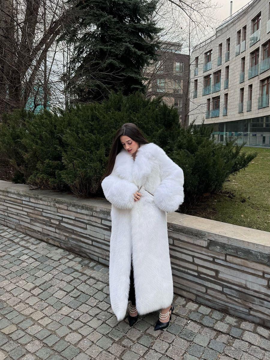 Woman in a full-length white faux fur coat, adjusting the lapels, standing on a cobblestone walkway.