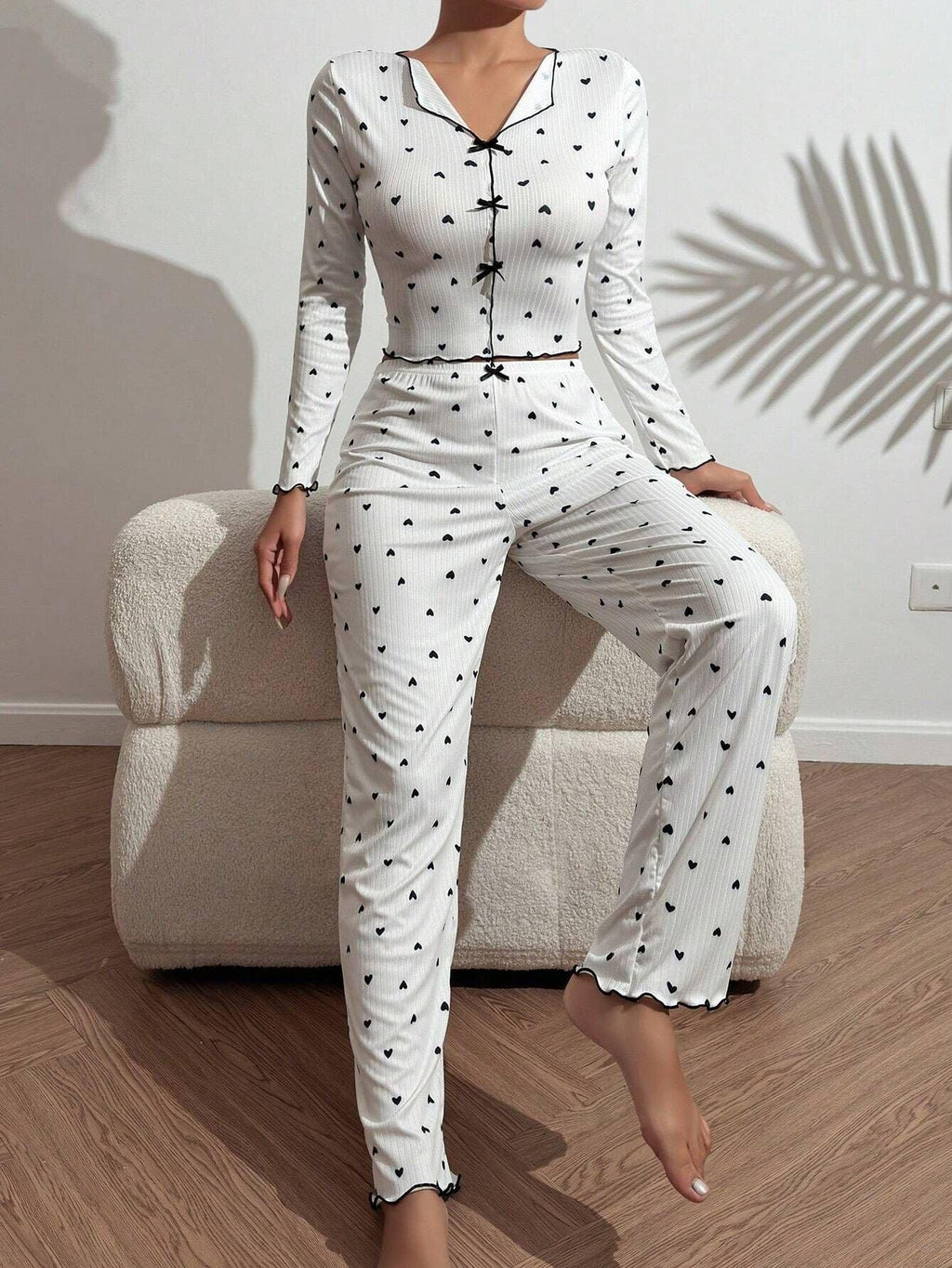 Woman wearing a white long-sleeve pajama set with a black heart print, sitting on a white armchair with one leg extended.