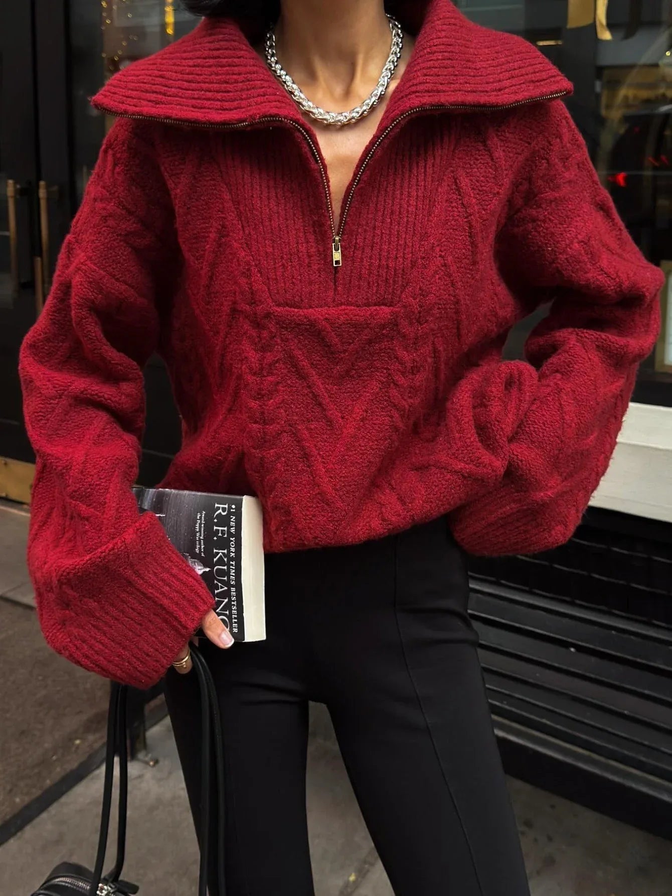 Woman wearing a red textured knit quarter zip sweater and black pants, holding a book and a handbag.