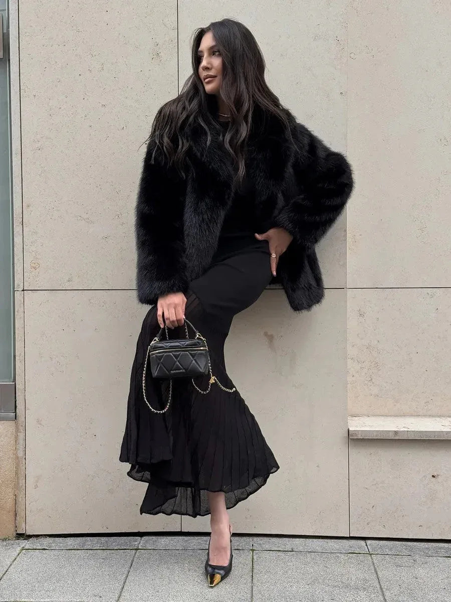 Woman wearing a black faux fur jacket over a black dress, leaning against a wall.