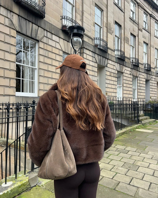 Back view of a woman wearing a brown faux fur cropped jacket, white crop top, and brown leggings, with a brown suede tote bag.